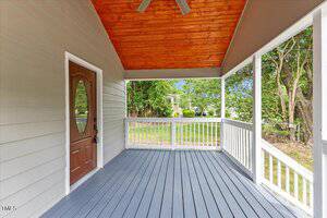 A covered front porch with gray floorboards, white railings, a wooden ceiling with a ceiling fan, and a brown door with an oval window, overlooking a grassy yard with trees.