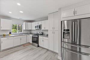 Modern kitchen with white cabinets, stainless steel appliances including a French door refrigerator, electric stove, and microwave, gray countertops, and wood-style flooring. A window above the sink provides natural light.