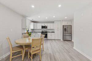 A modern kitchen with white cabinets, stainless steel appliances, and gray countertops. A round wooden dining table with four chairs and a vase of flowers sits on the light wood floor. The room is brightly lit.