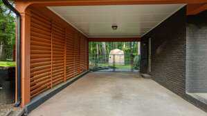 A covered carport with a concrete floor, orange support beams, slatted wood privacy screen on one side, black brick wall on the other, and a chain-link gate at the back opening to a yard with trees and a small shed.