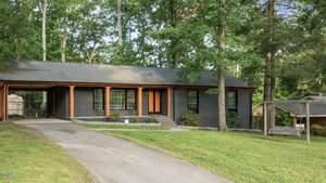 A single-story, dark gray brick house with a covered front porch, black-framed windows, and a sloped driveway. The yard is grassy with mature trees, and a wooden swing structure is on the right side.