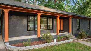 A single-story house with dark brick exterior, orange trim, black-framed windows, a covered front porch, and a small landscaped garden with shrubs and a concrete border.