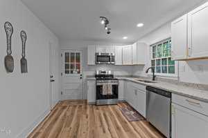 A modern kitchen with white cabinets, stainless steel appliances, light wood flooring, and granite countertops. There is a window above the sink and wall art shaped like a fork and spoon on the left wall.