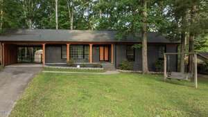 A single-story dark gray house at 307 Yowland Road, Henderson, features large windows, a covered carport on the left, a front porch with orange trim and steps, plus a wooden swing in the grassy front yard surrounded by trees.