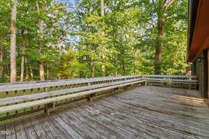 A large wooden deck with built-in benches is attached to a house, overlooking a wooded area with tall green trees. The weathered deck boards suggest outdoor use and exposure to the elements.