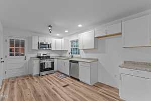 A modern kitchen with white cabinets, stainless steel appliances, granite countertops, wood flooring, a window above the sink, and a door leading outside. Ceiling lights and natural light brighten the room.