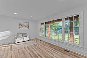 A bright living room with light wood flooring, a white loveseat, a beige rug, a piece of artwork on the wall, and large windows overlooking a green yard with trees and a wooden structure.