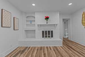 A white brick fireplace with a mantel holding a red potted plant is set against a white wall. Built-in shelves and two framed geometric artworks are on the left. The floor is light wood, and a gold leaf decor is on the right wall.