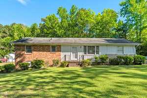 A charming single-story house at 1593 Washington Avenue in Creedmoor features a blend of brick and white siding, black roof, central door with steps, and is surrounded by lush green grass, bushes, and trees under a clear blue sky.