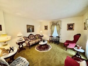 A living room with vintage furniture, including a patterned sofa, red armchair, wooden chairs, and ornate lamps. A small round table with a doily sits on a rug over green carpet. Framed art hangs on cream-colored walls.