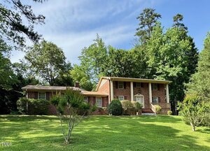 A two-story red brick house with white trim sits on a large, well-maintained lawn at 1818 Brookrun Road in Henderson. Mature trees and shrubs surround the home, which features a prominent front entrance with columns under a partly cloudy sky.