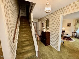 A hallway with patterned wallpaper, green carpeted stairs with a wooden handrail, a chandelier, a wooden dresser with a mirror, and a view into a living room with green carpet and classic furniture.
