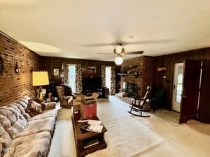 A spacious living room with brick and wood-paneled walls, a ceiling fan with a light, a sofa, armchairs, coffee table, boxes, a fireplace, TV, and a door leading outside. The room is carpeted and well-lit.