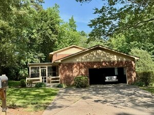 A brick house with a two-car garage, one door open showing a parked car inside. The home has a screened porch on the left and is surrounded by green trees and grass under a clear sky.