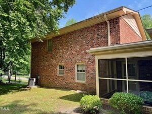 A two-story brick house with a side yard, visible air conditioning unit, screened porch, and shrubs in the foreground. Trees provide partial shade over the house. The sky is mostly clear.