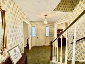 A hallway with patterned wallpaper, carpeted floor, white front door flanked by narrow windows, a staircase with a white railing, a framed photo on a table, and a hanging light fixture on the ceiling.