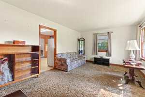A living room with a patterned sofa, a wooden bookshelf with a doll, a grandfather clock, a small black TV stand under a window, a lamp on a side table, and carpeted flooring. Sunlight streams through the windows.