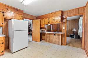 Kitchen with wood-paneled walls and cabinets, white refrigerator, open door leading to a living room with carpet, and a counter with a lower serving area. Fluorescent ceiling light is on.
