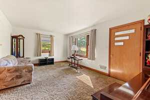 A modest living room with beige carpet, a floral sofa, a grandfather clock, two windows with white curtains, a wooden door, a small black TV stand, a side table with a lamp, and natural light entering the room.