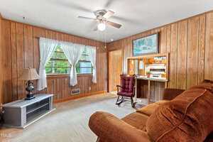A living room with wood-paneled walls, beige carpet, a brown sofa, a red cushioned rocking chair, a small table with a lamp, and two windows with sheer white curtains. A ceiling fan and pass-through to kitchen are visible.