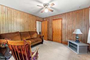 A living room with wood-paneled walls, a brown upholstered sofa, a wooden rocking chair, a ceiling fan with a light, beige carpet, two wooden doors, and a TV stand with a lamp by a window with white curtains.