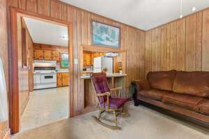 A living room with wood-paneled walls, a brown couch, a maroon rocking chair, and a pass-through counter to a kitchen with wooden cabinets, a white stove, and a refrigerator. A painting hangs above the counter.