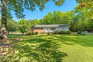 A single-story brick and white siding house sits on a large, well-maintained lawn with green grass and mature trees under a clear blue sky. Shrubs line the front of the house.