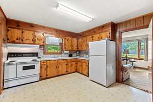 A kitchen with wood-paneled cabinets, white appliances including a stove and refrigerator, a window above the sink with a valance, and adjoining view of a living room with a large window.