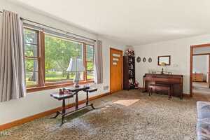 A living room with large windows, beige carpet, a wooden table with a lamp, a piano against the wall, and several decorative plates and dolls on display; an open doorway leads to another room.