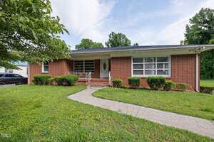 Single-story red brick house with a covered front porch, white trim, and a light-colored front door. A concrete walkway leads to the entrance, with neatly trimmed bushes and a grassy lawn in front.