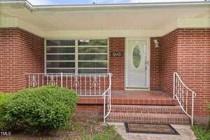 A red brick house with a covered front porch, white railing, a glass panel front door, and a "Home Sweet Home" doormat. There is a bush next to the brick steps and a window with horizontal blinds.