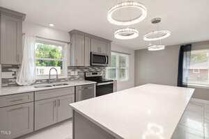 Modern kitchen at 2315 Oxford Road, Henderson features gray cabinets, stainless steel appliances, white marble countertops, a large island, tile backsplash, and three circular pendant lights. Large windows provide ample natural light.