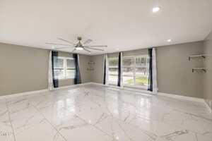 A spacious, empty room with large white marble floor tiles, gray walls, two large windows with dark curtains, a ceiling fan with lights, and wall-mounted shelves on either side.
