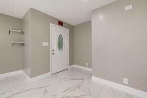 A small entryway with light gray walls, white crown molding, large white marble tile flooring, a white door with an oval window, two black wall shelves, and an exit sign above the door.