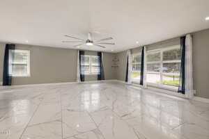 Spacious empty room with large white marble floor tiles, gray walls, a ceiling fan with metal blades, several windows with blue and white curtains, and recessed lighting in the ceiling.