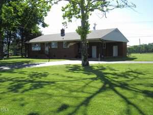 A one-story brick house with a gray roof sits in a large, well-maintained yard. A tree with green leaves casts a shadow on the grass in the foreground. A concrete driveway leads to the house.