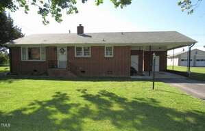 Charming single-story red brick house at 1700 Warrenton Road in Henderson features a gray roof, small front steps, covered carport on the right, and spacious green lawn shaded by a tree. A detached garage sits in the background.