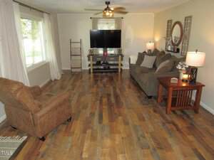 A living room with wood flooring, a brown armchair, a sofa, a wooden side table with two lamps, a TV on a stand, a ceiling fan, and large windows with light curtains. Neutral-colored walls and decor.