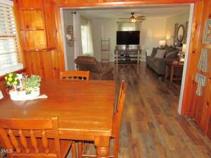 A dining area with a wooden table and chairs opens into a living room with a sofa, armchair, TV, and wooden floors. The walls are wood-paneled in the dining area and light-colored in the living room.