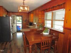 A kitchen with wood-paneled walls and cabinets, a wooden dining table with four chairs, stainless steel appliances, a window with white blinds, and a ceiling light fixture. The door is open to the outside.