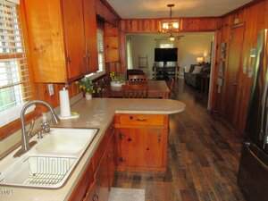 A kitchen with wood-paneled cabinets and walls, a double sink, and beige countertops. The space leads to a dining area with a wooden table and a living room with a TV and a couch in the background.