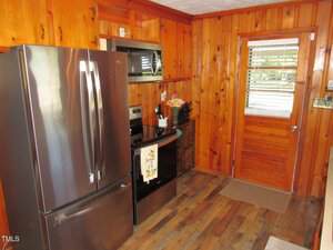 Kitchen with wood-paneled walls and wood flooring, featuring a stainless steel refrigerator, oven, microwave, and a wooden door with a window and white blinds, as well as a small mat on the floor.