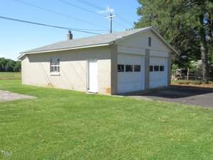 A single-story, detached garage with two white garage doors, light-colored cinder block walls, a gray shingle roof, and a window on the side, surrounded by grass and trees in the background.