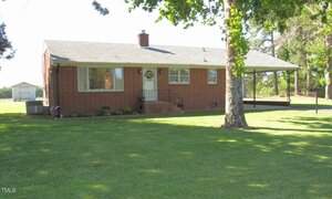 Charming single-story red brick house at 1700 Warrenton Road in Henderson, featuring a gray shingle roof, central chimney, small front porch, large grassy lawn, tree in the foreground, and a covered carport on the right.