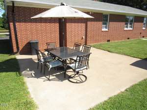 A rectangular black patio table with six metal chairs and a beige umbrella is set on a concrete patio next to a red brick house with green grass on both sides.