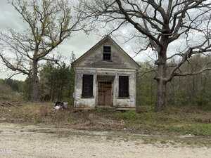 A small, old, white wooden house with boarded-up windows and a decaying exterior stands alone beside a dirt road. Overgrown grass and trees surround the abandoned structure under a cloudy sky.