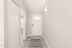 A narrow hallway with light gray walls and a white front door, featuring a small rug on the floor and a closet door on the left. The space is well-lit with a wall-mounted light fixture.