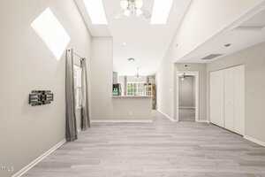A bright, empty living space with light gray wood flooring, light gray walls, a ceiling fan with lights, a wall-mounted TV bracket, large windows, skylights, and an open view into a kitchen with white cabinetry.