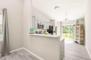 A modern kitchen with light gray cabinets, stainless steel appliances, and a white countertop, opening to a bright living area with large windows and a wooden cabinet near sliding glass doors.