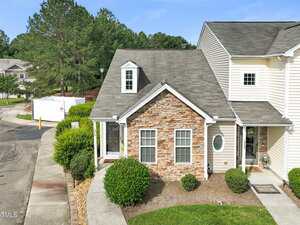 A two-story townhouse with a stone and beige siding exterior, small front porch, shrubs, and a manicured lawn on a corner lot, with a sidewalk and a street curving around the property.